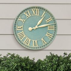 a clock on the side of a building with bushes around it and trees in front of it