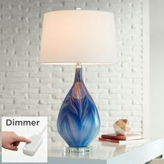 a blue glass lamp on a table with a white shade over it and a hand pointing at the light