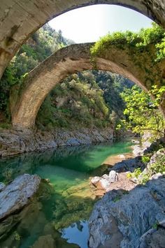 an old stone bridge over a river surrounded by trees and rocks, with water running under it