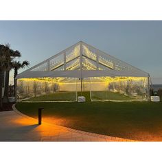 a large white tent is lit up at night with lights on the grass and palm trees in the background