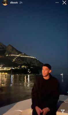 a man sitting on top of a boat in front of a mountain and water at night