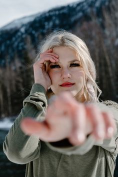 a blonde woman holding her hand near her face and pointing at the camera with mountains in the background