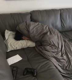 a person sleeping on top of a couch covered by a blanket and pillows with video game remotes