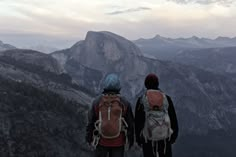 two people with backpacks standing on top of a mountain looking out at the mountains