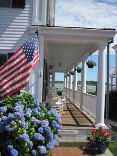 an american flag on the porch of a white house with blue hydrangeas in front