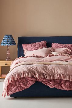 a bed with pink ruffled sheets and pillows on it, next to a night stand