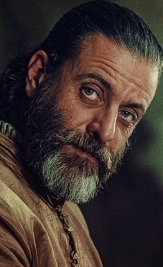 a close up of a man with a beard and wearing a brown shirt looking at the camera