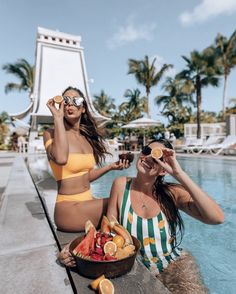 two women in bathing suits sitting on the edge of a swimming pool with fruit and drinks