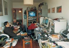 two men sitting in chairs working on their laptops at the same time, while another man is watching television