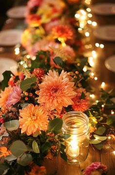 an arrangement of flowers and candles on a long table with lights in the back ground