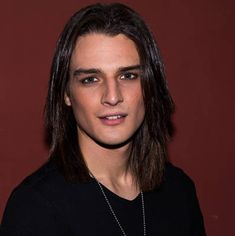 a young man with long hair wearing a black shirt and silver necklace, standing in front of a red wall