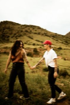 two people holding hands while walking through a field with hills in the background and one person wearing a red hat