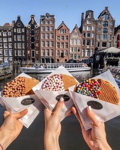 three people are holding up their food in front of some buildings on the water with boats in the background