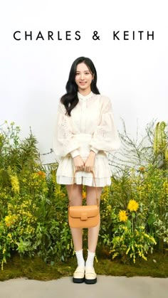 a woman in white holding a tan purse and posing for the camera with yellow flowers behind her