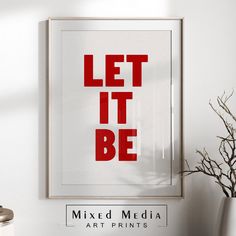 a red print that says let it be on the wall next to a white vase