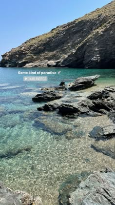 some clear blue water and rocks on the shore with an island in the background that says some kind of paradise