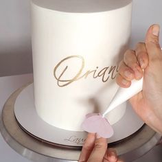 a woman is decorating a white cake with gold lettering on it and a pink flower