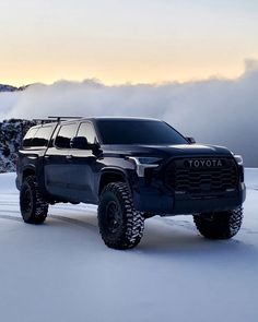 a black truck is driving through the snow in front of some mountains and clouds on a cloudy day