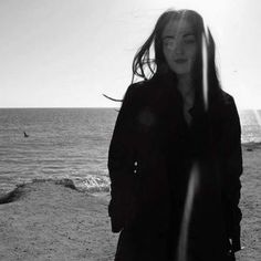 a woman standing on top of a sandy beach next to the ocean with her hair blowing in the wind