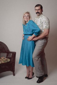 a man and woman standing next to each other in front of a white wall with a wicker chair