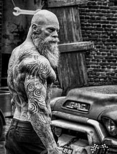 a man with tattoos on his arms and chest standing next to a motorcycle in front of a brick building