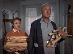 an older man standing next to a young woman holding stacks of wood in her hands