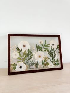 a wooden frame with white flowers and green leaves in it on top of a table