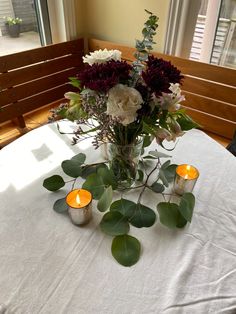 a vase with flowers and candles on a white table cloth in front of a window