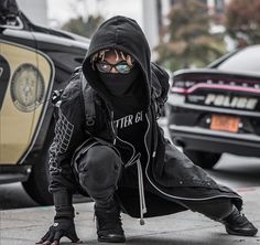 a person in a black hoodie crouching down on the ground next to a parked car