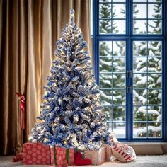 a blue christmas tree in front of a window with presents on the floor next to it