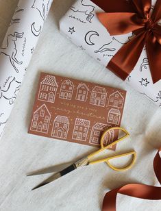 a pair of scissors sitting next to some wrapping paper with houses on it and red ribbon