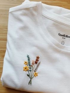 a white t - shirt with flowers embroidered on the front and side, sitting on a wooden surface