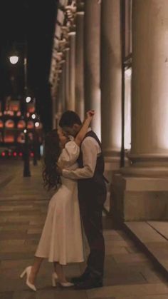 a man and woman standing next to each other on a sidewalk at night with lights in the background