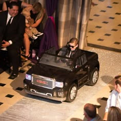 a man in a suit driving a black truck down a runway with people watching from the sidelines