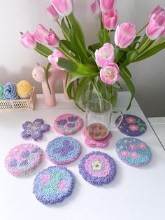 pink and purple crocheted coasters sitting on a table next to a vase filled with tulips