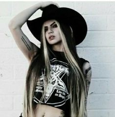 a woman with long hair wearing a cowboy hat and black shirt standing in front of a white brick wall