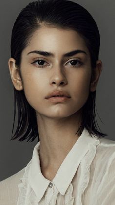 a woman with black hair wearing a white shirt and looking at the camera while standing in front of a gray background