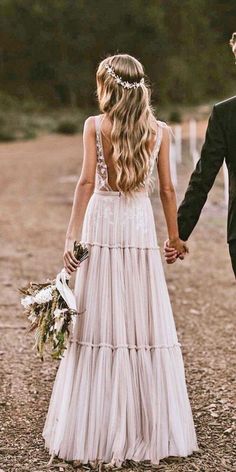 a man and woman holding hands walking down a dirt road with trees in the background