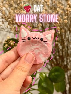 a hand holding up a pink cat brooch that says worry stone on it's face