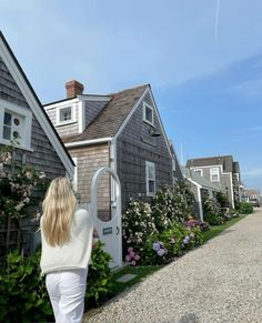 a woman walking down a gravel road next to a gray house with white shutters