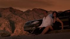 a man and woman sitting next to a car in front of mountains at sunset with the sun setting