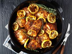 chicken with lemons and herbs in a skillet on a wooden table next to utensils