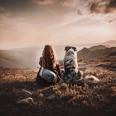 a woman sitting on top of a grass covered field next to a dog in the mountains