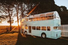 a white double decker bus parked in the grass next to some trees and a fence