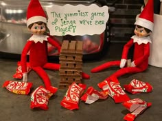 two elfs are sitting next to a pile of candy and one is holding a sign