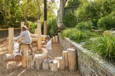 two children are playing in an outdoor play area with wooden logs and rocks, while another child is on the other side of the fence