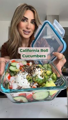 a woman holding up a large glass bowl filled with vegetables and cucumber slices