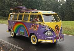 an old vw bus painted in rainbow colors driving down the road with trees and grass behind it