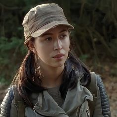 a woman wearing a hat and jacket standing in the dirt with trees in the background