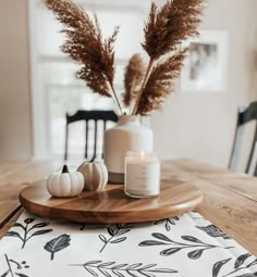 a wooden table topped with a white vase filled with dried plants and small pumpkins